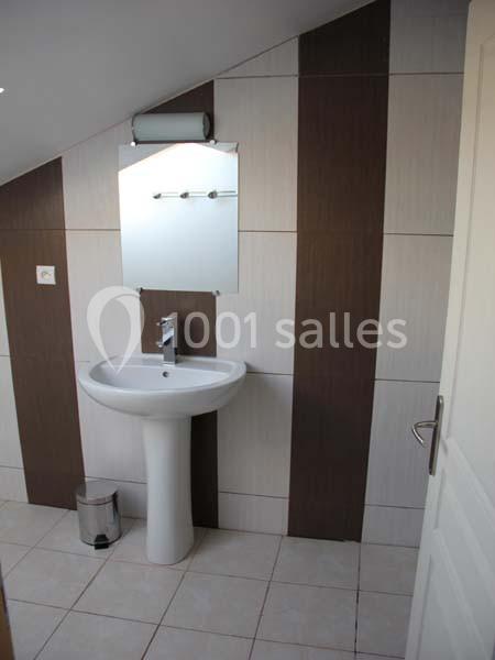 Lavabo blanc avec miroir rectangulaire, éclairage mural et carrelage beige et marron dans une salle de bain mansardée.