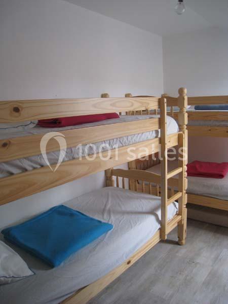 Lits superposés en bois clair avec draps blancs, couvertures pliées rouges et bleues dans une pièce lumineuse.