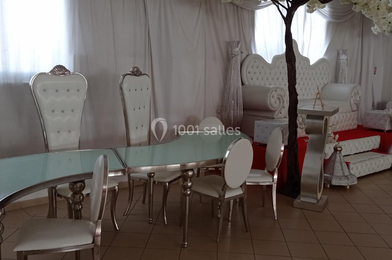 Salle de réception avec mobilier blanc et argenté, incluant des fauteuils capitonnés, une table en verre et des décorations…
