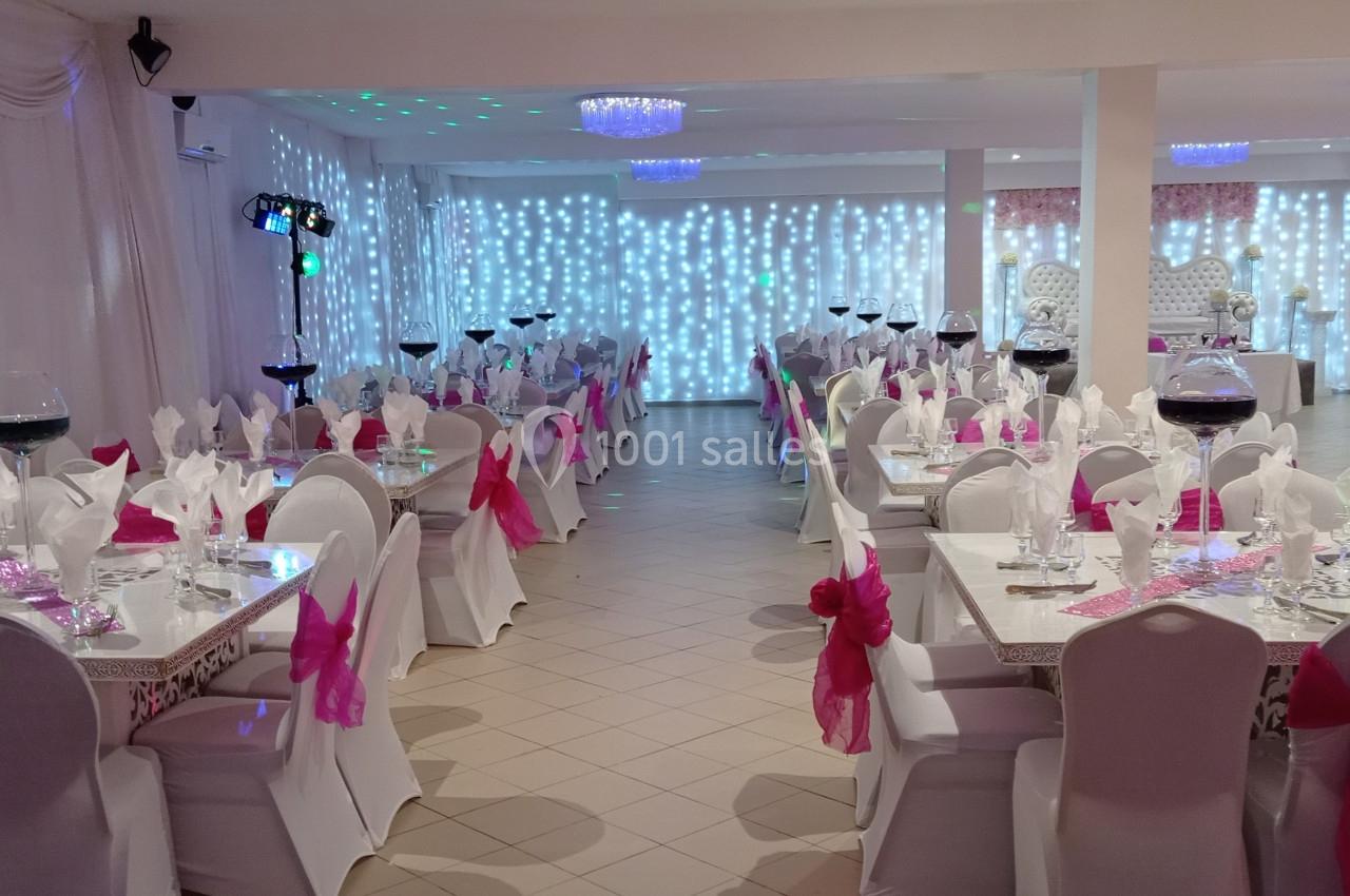 Salle de réception décorée avec des tables dressées, nappes blanches, nœuds roses et éclairage lumineux en arrière-plan.