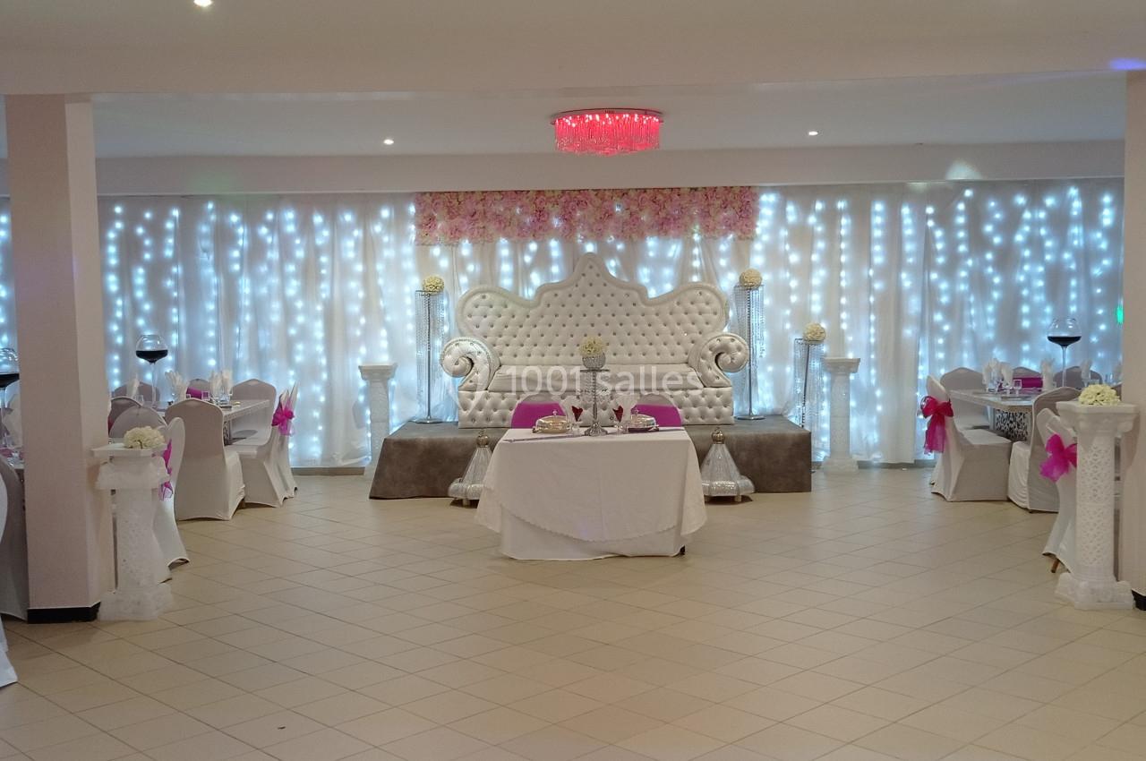 Salle de réception décorée avec des tables rondes, des chaises ornées de nœuds roses et un canapé blanc sur une estrade.