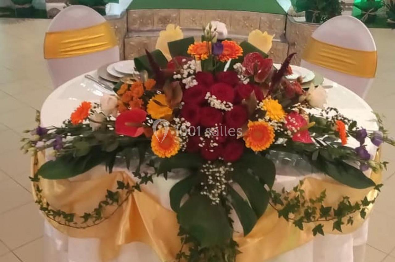 Table décorée avec des fleurs colorées et rubans dorés, devant une estrade ornée d'un canapé blanc capitonné.