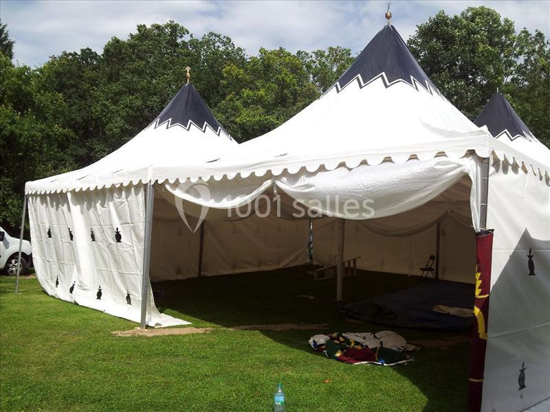 Tente blanche à deux pointes installée sur une pelouse, entourée d'arbres, avec des objets posés au sol.