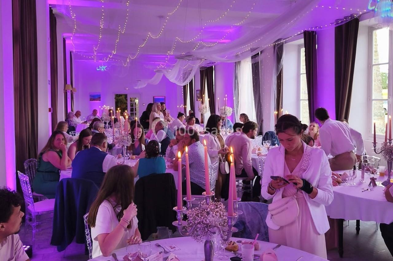 Salle de réception décorée avec des guirlandes lumineuses, des convives assis et debout lors d'un événement festif.