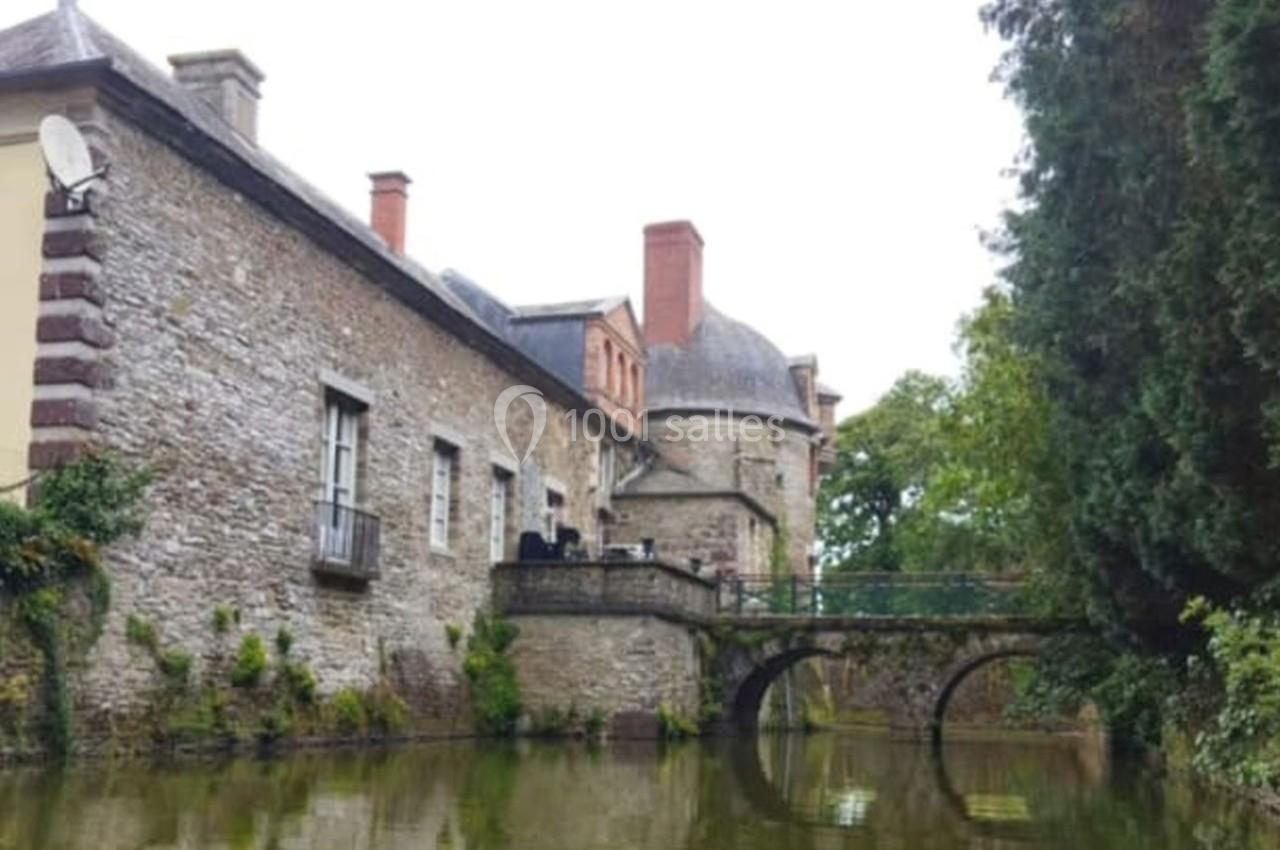 Vue d'un château en pierre avec un pont en arc traversant un fossé rempli d'eau, entouré de végétation.