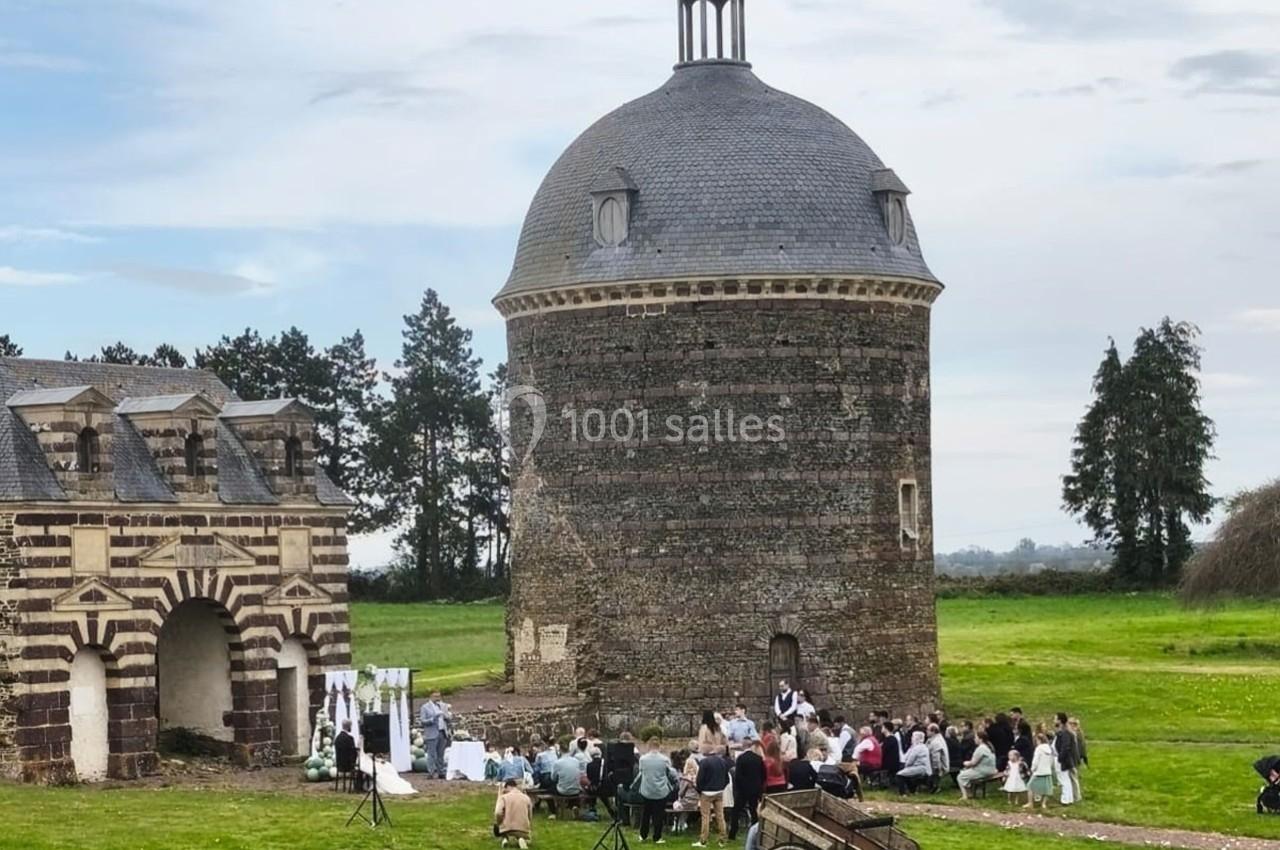 Cérémonie en plein air avec des invités assis devant une tour en pierre et un bâtiment ancien, dans un cadre champêtre.