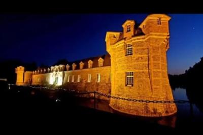 Château illuminé de nuit avec des murs en pierre, entouré d'arbres et d'une chaîne métallique au premier plan.