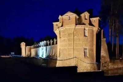 Château illuminé de nuit avec des murs en pierre, entouré d'arbres et d'une chaîne métallique au premier plan.