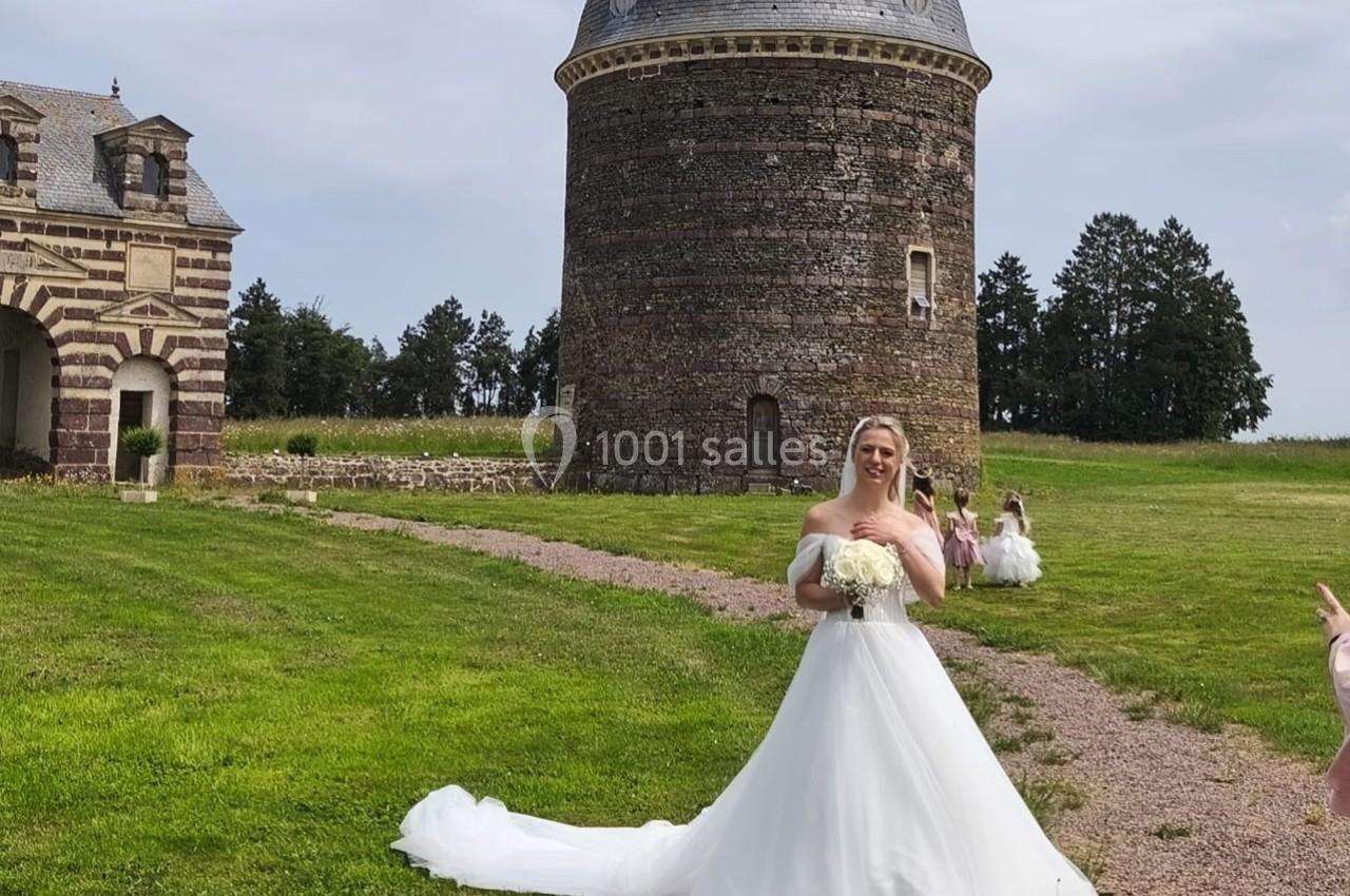 Une mariée en robe blanche pose devant une tour en pierre et un bâtiment ancien, sur une pelouse verdoyante.