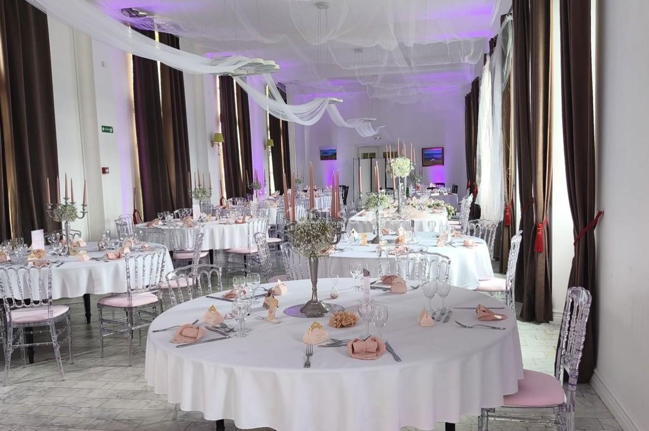 Salle de réception décorée avec des tables rondes dressées, nappes blanches, chandeliers et éclairage violet.