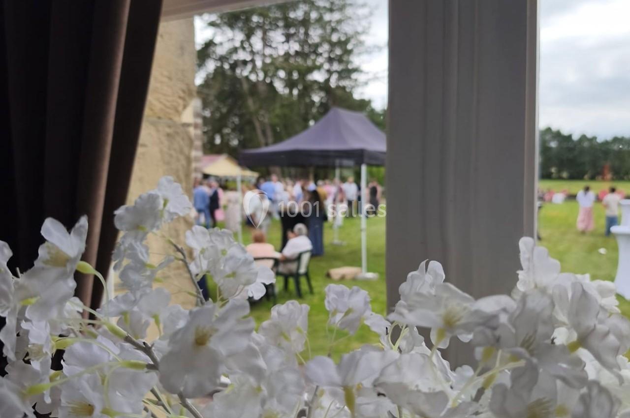 Vue depuis une fenêtre sur un jardin avec des fleurs blanches au premier plan et des personnes rassemblées sous une tente.