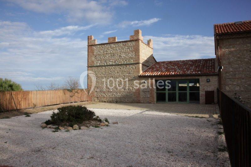 Bâtiment en pierre avec une tour crénelée, cour gravillonnée et ciel partiellement nuageux en arrière-plan.