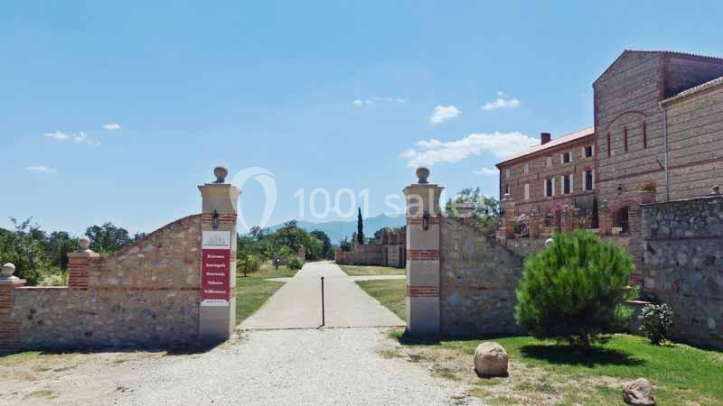 Entrée d'une propriété avec portail ouvert, allée centrale, bâtiments en pierre et paysage montagneux en arrière-plan.