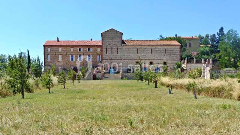 Bâtiment en pierre avec toit en tuiles rouges, entouré d'un champ et de quelques arbres sous un ciel dégagé.