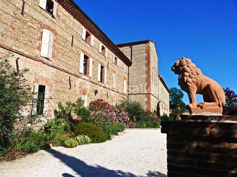 Bâtiment en pierre avec volets blancs, jardin fleuri et statue de lion en terre cuite sur un muret au premier plan.