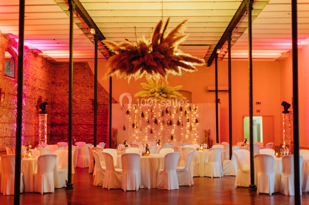 Salle de réception décorée avec des guirlandes lumineuses, des tables rondes dressées et des chaises blanches.