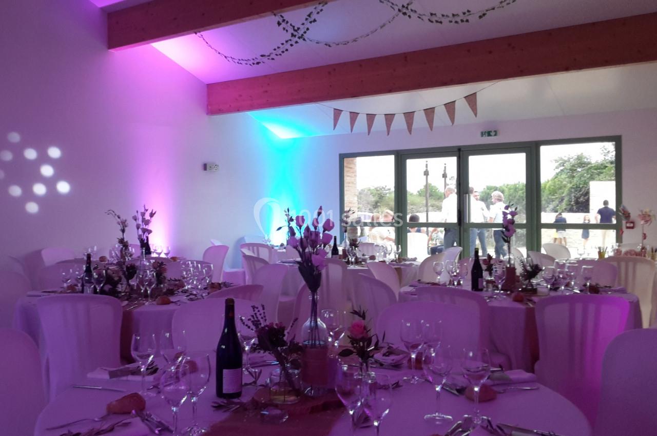 Salle de réception décorée pour un événement, avec tables dressées, nappes blanches et éclairage coloré.