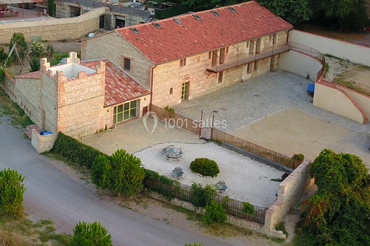 Vue aérienne d'une maison en pierre avec toit en tuiles, cour pavée, jardin clôturé et arbres environnants.
