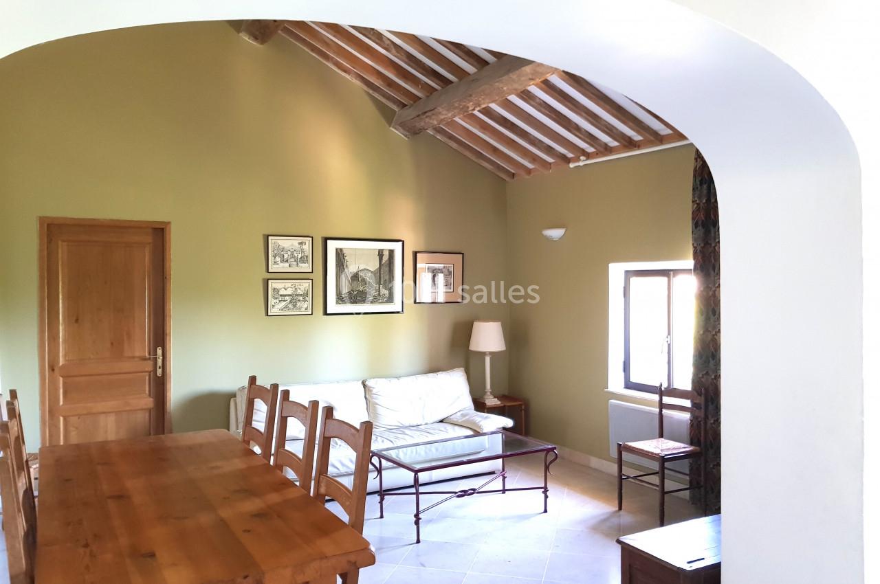 Salle de séjour lumineuse avec table en bois, chaises, canapé, lampadaire, cadres au mur et poutres apparentes.