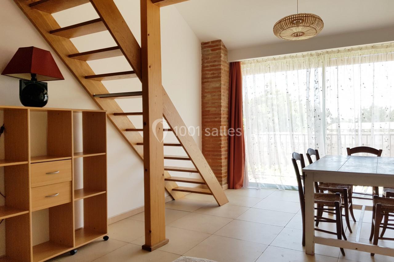 Escalier en bois dans un salon lumineux avec table, chaises, étagère et grande baie vitrée donnant sur l'extérieur.