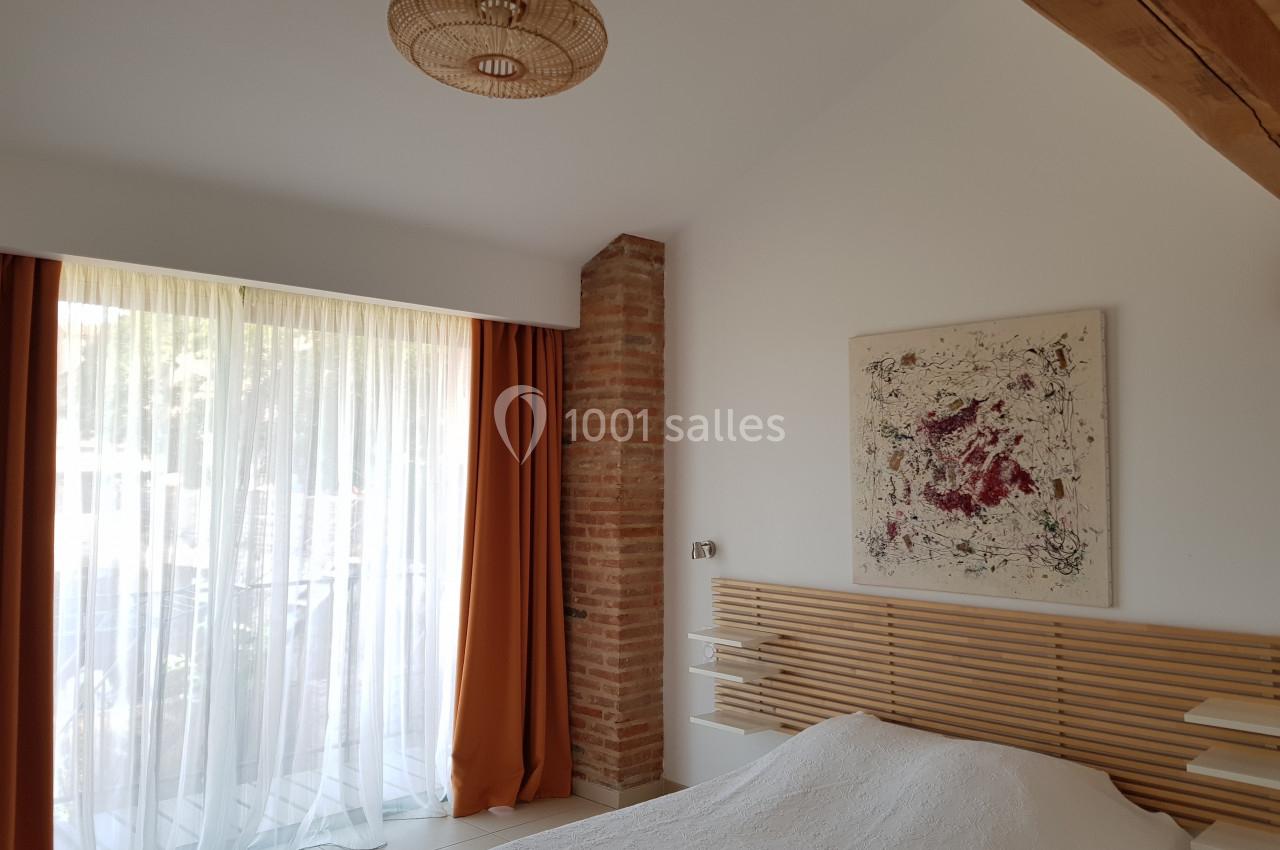 Chambre lumineuse avec lit, tête de lit en bois, tableau abstrait, rideaux orange et mur en briques apparentes.