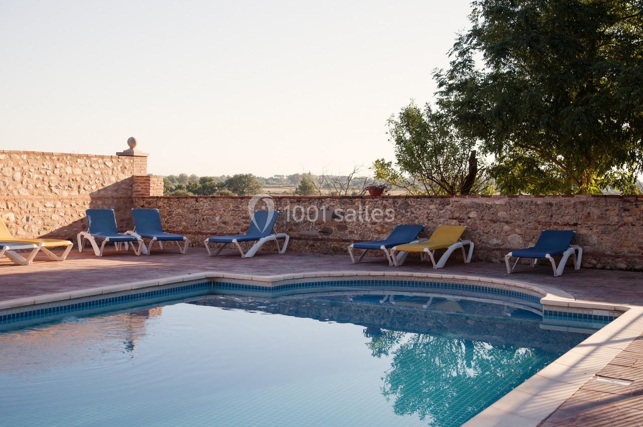 Piscine extérieure entourée de chaises longues colorées, avec un mur en pierre et des arbres en arrière-plan.
