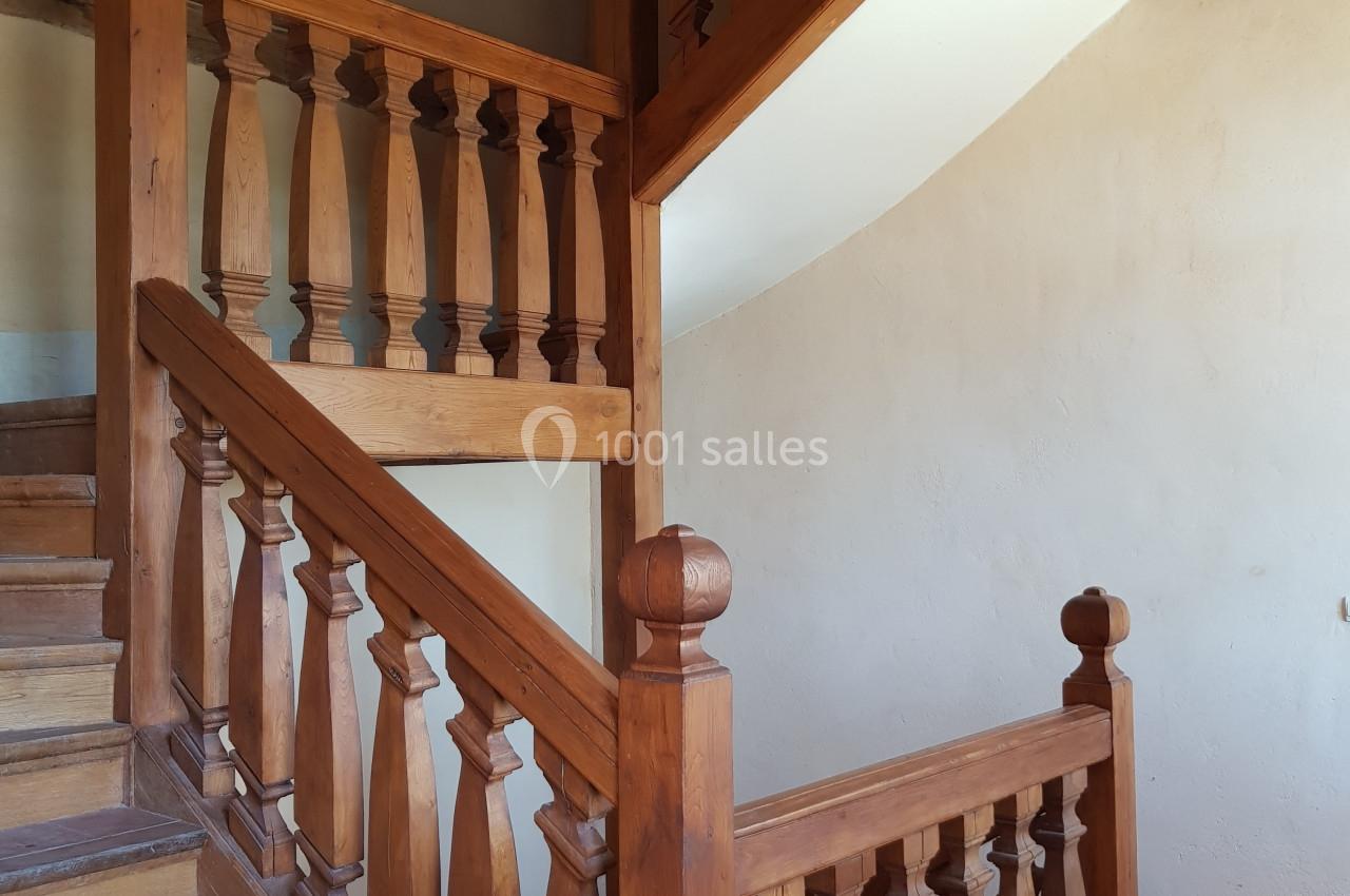 Escalier en bois avec balustrades sculptées, situé dans un intérieur aux murs clairs et poutres apparentes.