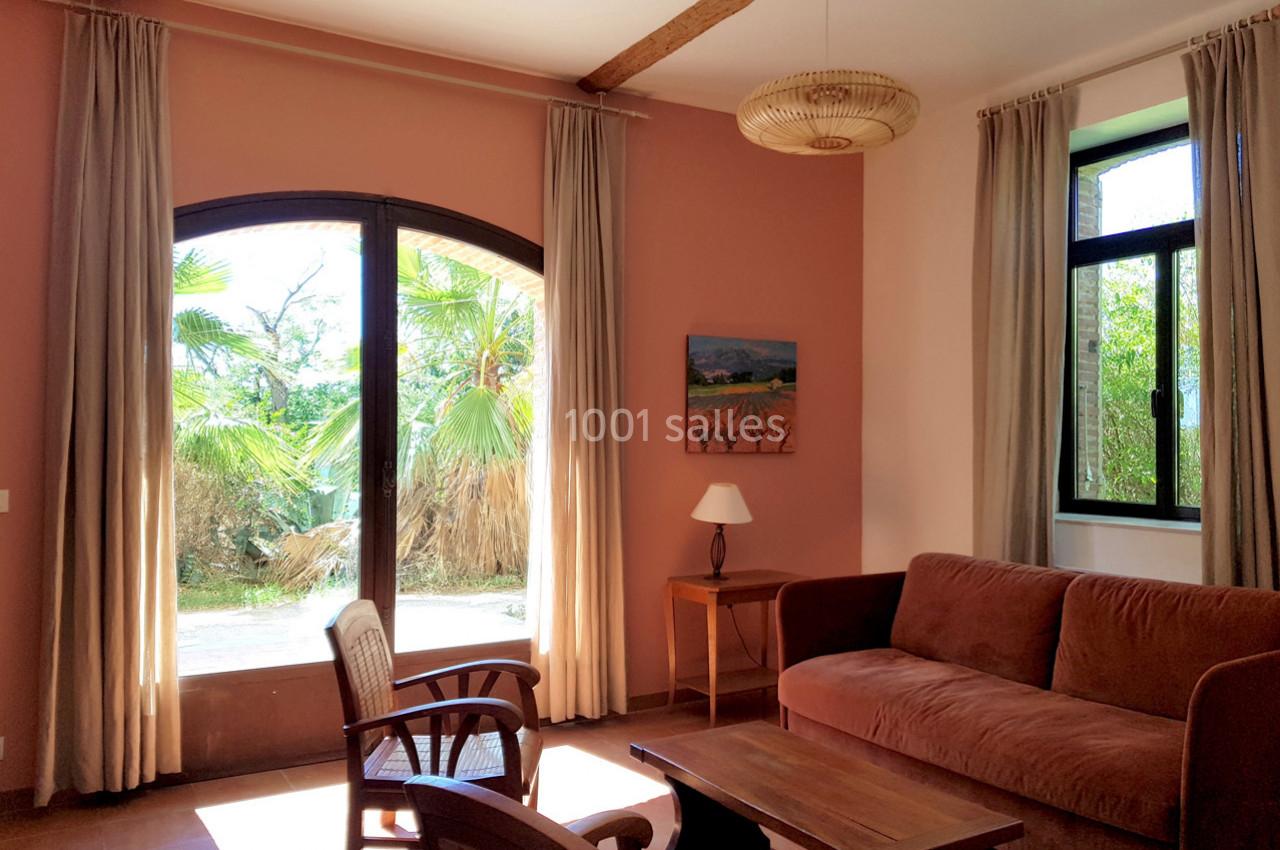 Salon lumineux avec murs roses, canapé, fauteuils, table basse, grande baie vitrée et vue sur jardin verdoyant.