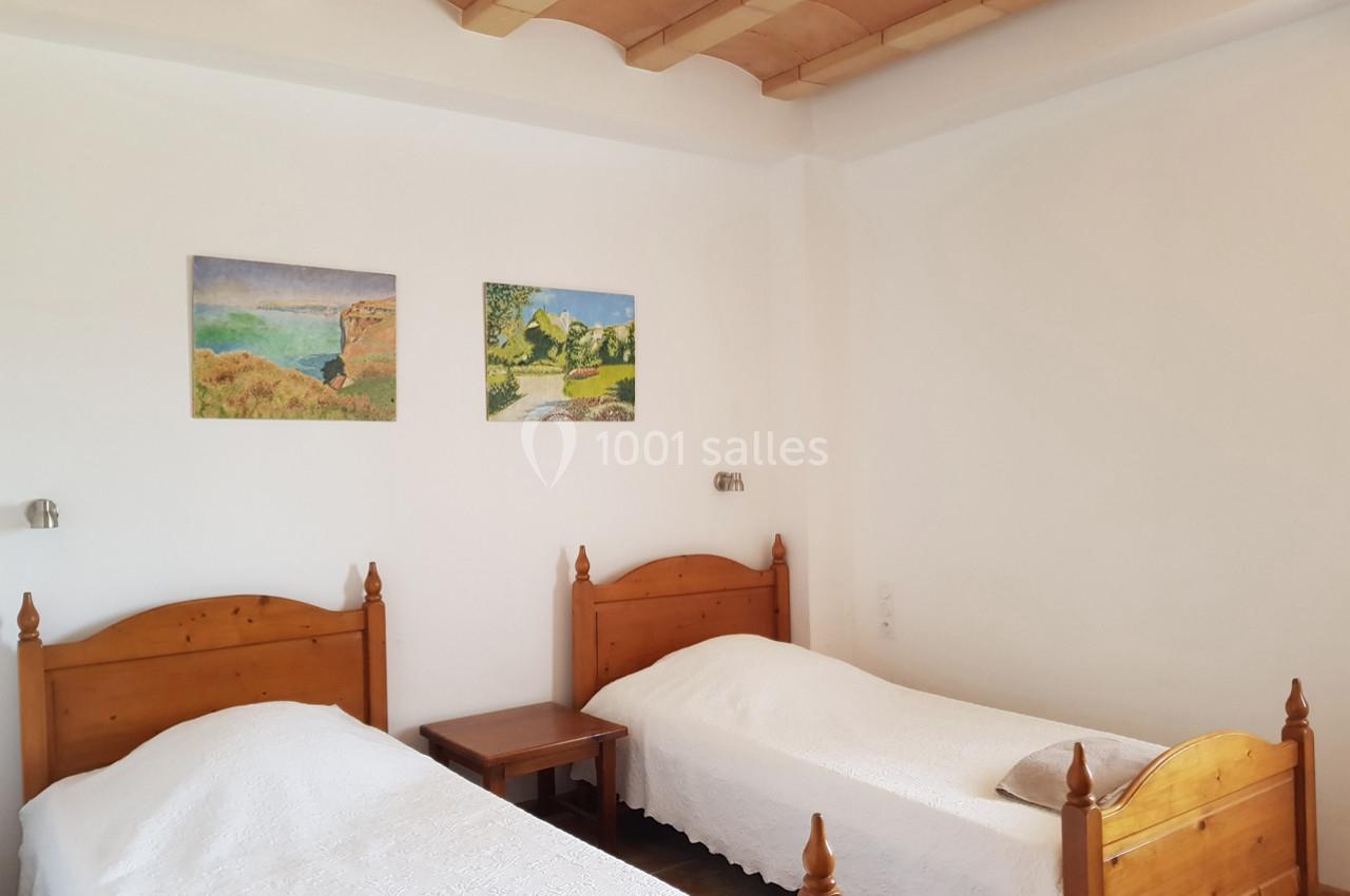 Deux lits simples en bois avec des couvre-lits blancs, une table de chevet et des tableaux accrochés sur un mur blanc.