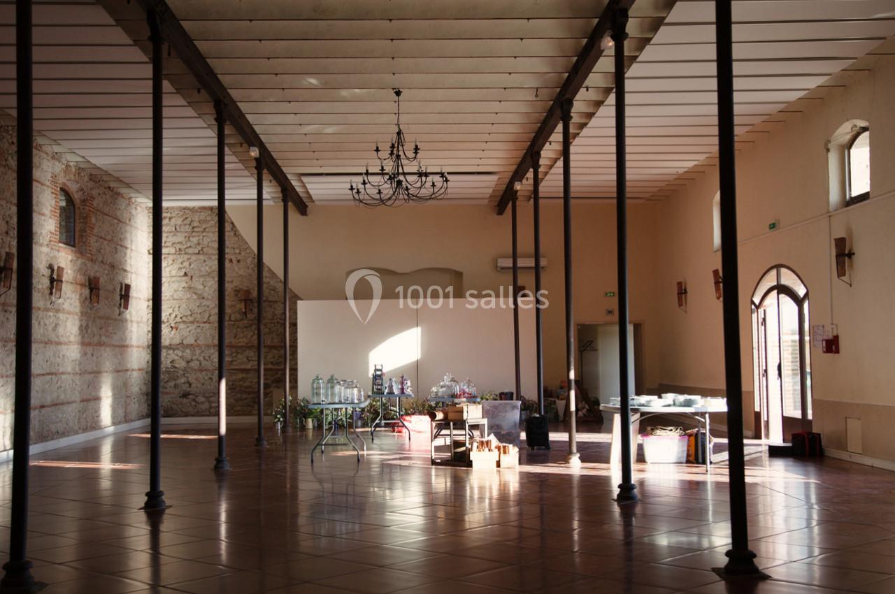 Grande salle lumineuse avec murs en pierre, poutres apparentes, sol carrelé et mobilier dispersé au centre.