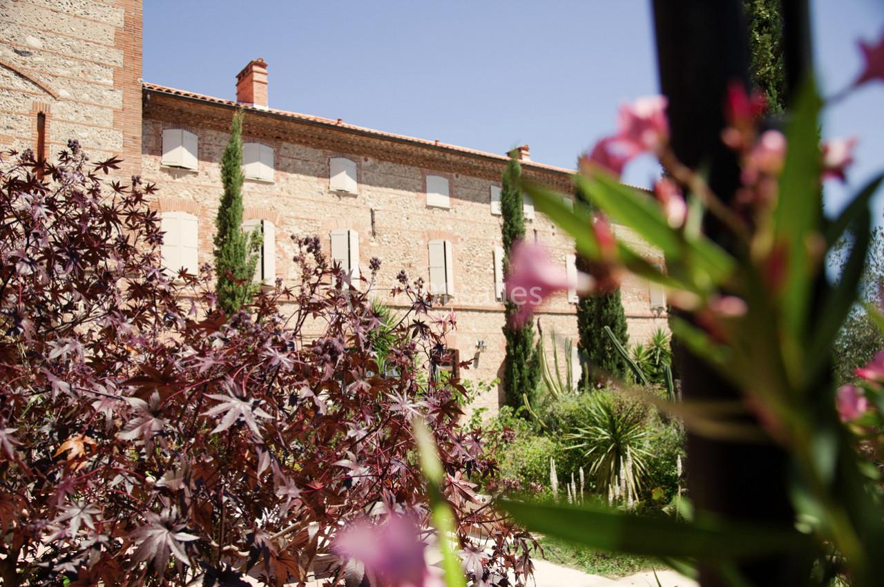 Façade en pierre d'un bâtiment entouré de végétation, avec des arbustes et des fleurs au premier plan.