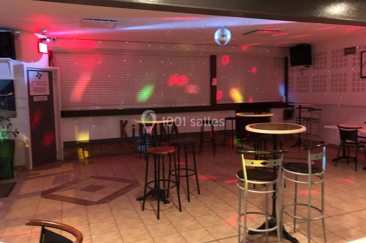 Salle vide avec tables hautes, chaises et éclairage coloré diffus, ambiance de soirée dans un espace carrelé.