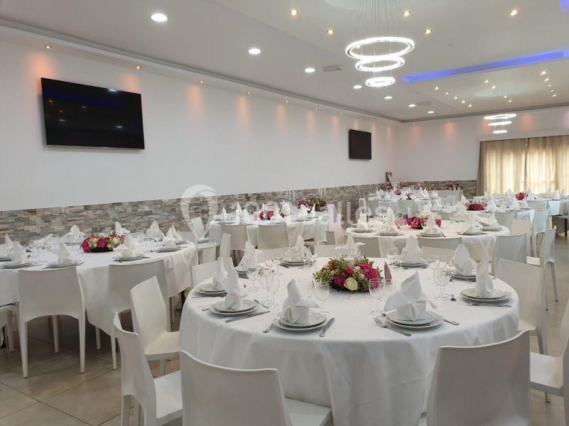 Salle de réception lumineuse avec tables rondes dressées, nappes blanches et décorations florales au centre.