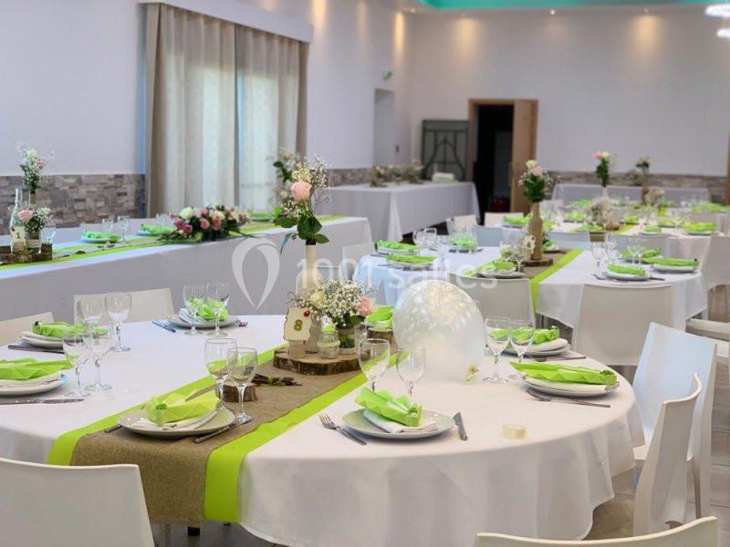 Salle de réception décorée avec des tables rondes, nappes blanches, chemins de table verts et arrangements floraux.
