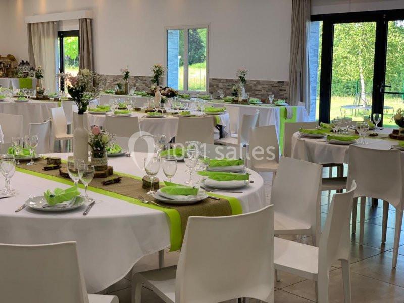 Salle de réception lumineuse avec tables rondes dressées, nappes blanches et décorations vertes et florales.