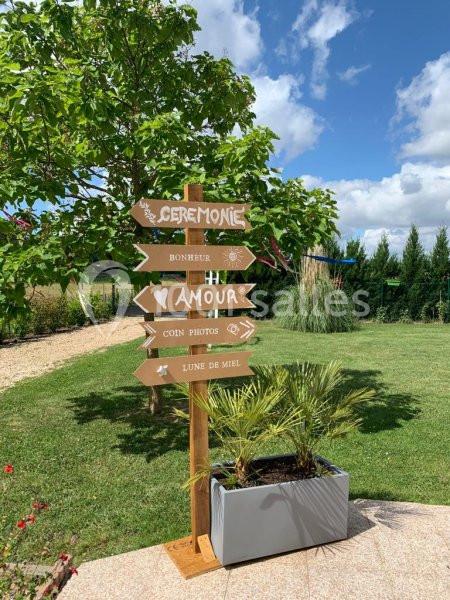 Panneau en bois avec des flèches indiquant ’Cérémonie’, ’Bonheur’, ’Amour’, ’Coin photos’ et ’Lune de miel’ dans un jardin.