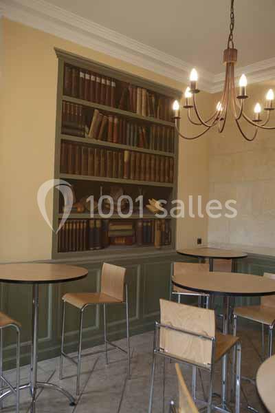 Salle avec des tables hautes, chaises en bois, bibliothèque remplie de livres anciens et lustre suspendu.