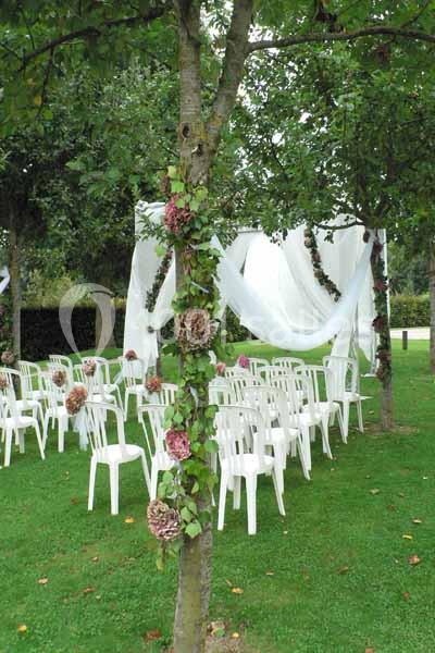 Chaises blanches disposées en extérieur sous des arbres décorés de fleurs et de voiles blancs.