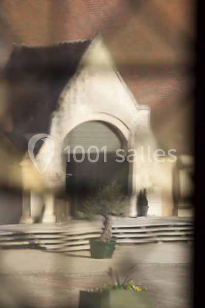 Entrée d'un bâtiment en pierre avec une arche, vue à travers une surface floue, un pot de fleurs au premier plan.