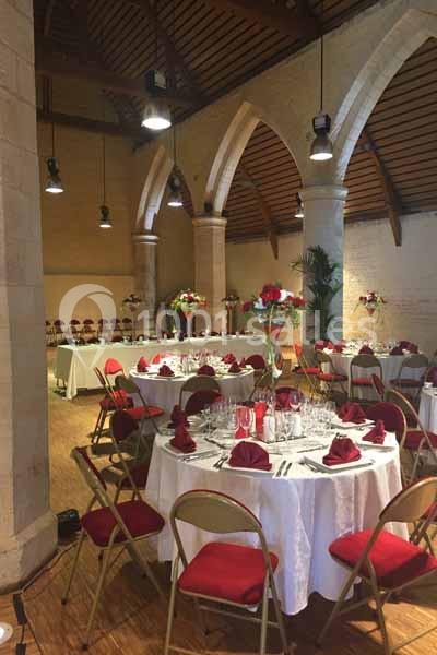 Salle de réception avec tables rondes dressées, nappes blanches, serviettes rouges et arches en pierre apparente.