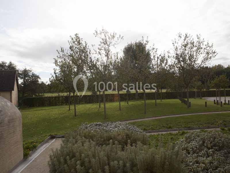 Un jardin avec des arbres alignés, des haies et des parterres de plantes sous un ciel légèrement nuageux.
