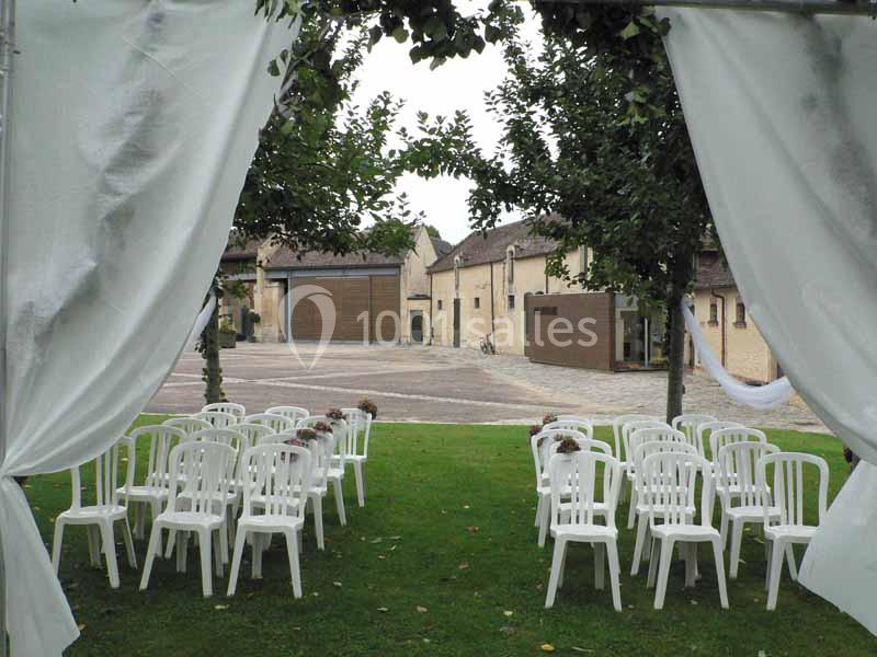 Chaises blanches disposées en rangées sur une pelouse, encadrées par des rideaux blancs, avec des bâtiments en arrière-plan.