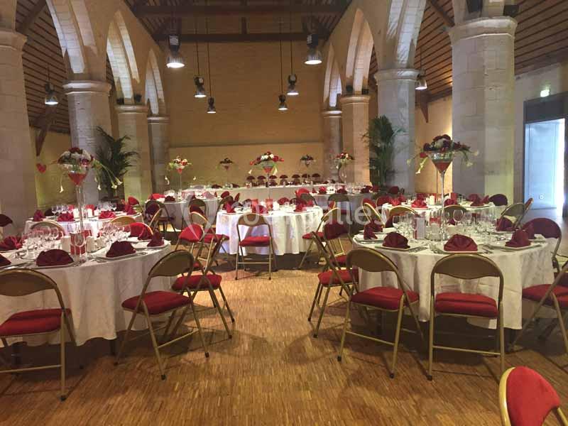 Salle de réception avec tables rondes dressées, nappes blanches, chaises rouges et décorations florales.