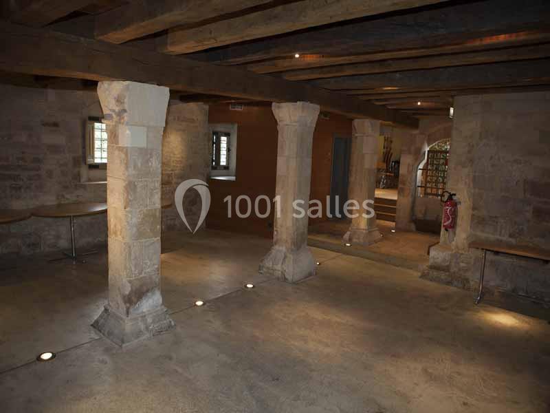 Salle en pierre avec poutres apparentes, colonnes et éclairage au sol, donnant sur une autre pièce par une ouverture.