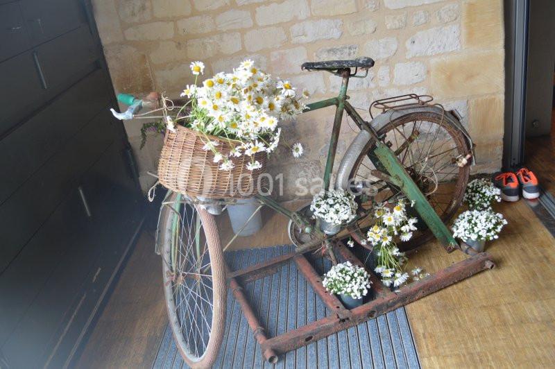 Vélo ancien décoré de paniers remplis de marguerites blanches, posé sur un tapis dans un intérieur en pierre.