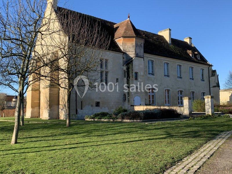 Bâtiment ancien en pierre avec toit en tuiles, entouré d'un jardin et d'arbres dénudés sous un ciel bleu.