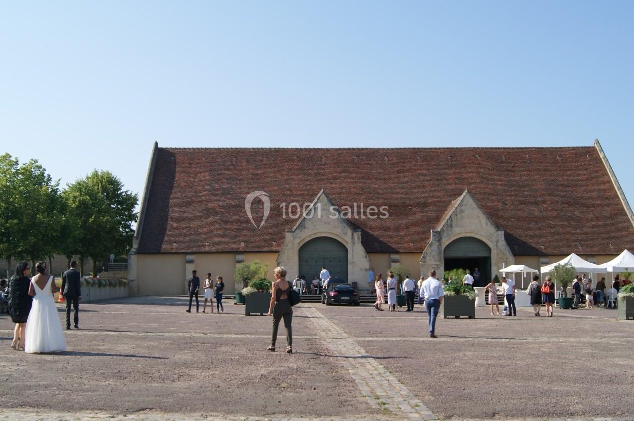 Grande cour pavée devant un bâtiment ancien avec toit en tuiles, des personnes rassemblées et des arbres en arrière-plan.