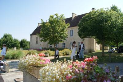 Un bâtiment en pierre avec un toit en tuiles rouges, entouré de jardins fleuris, accueille un groupe de personnes.