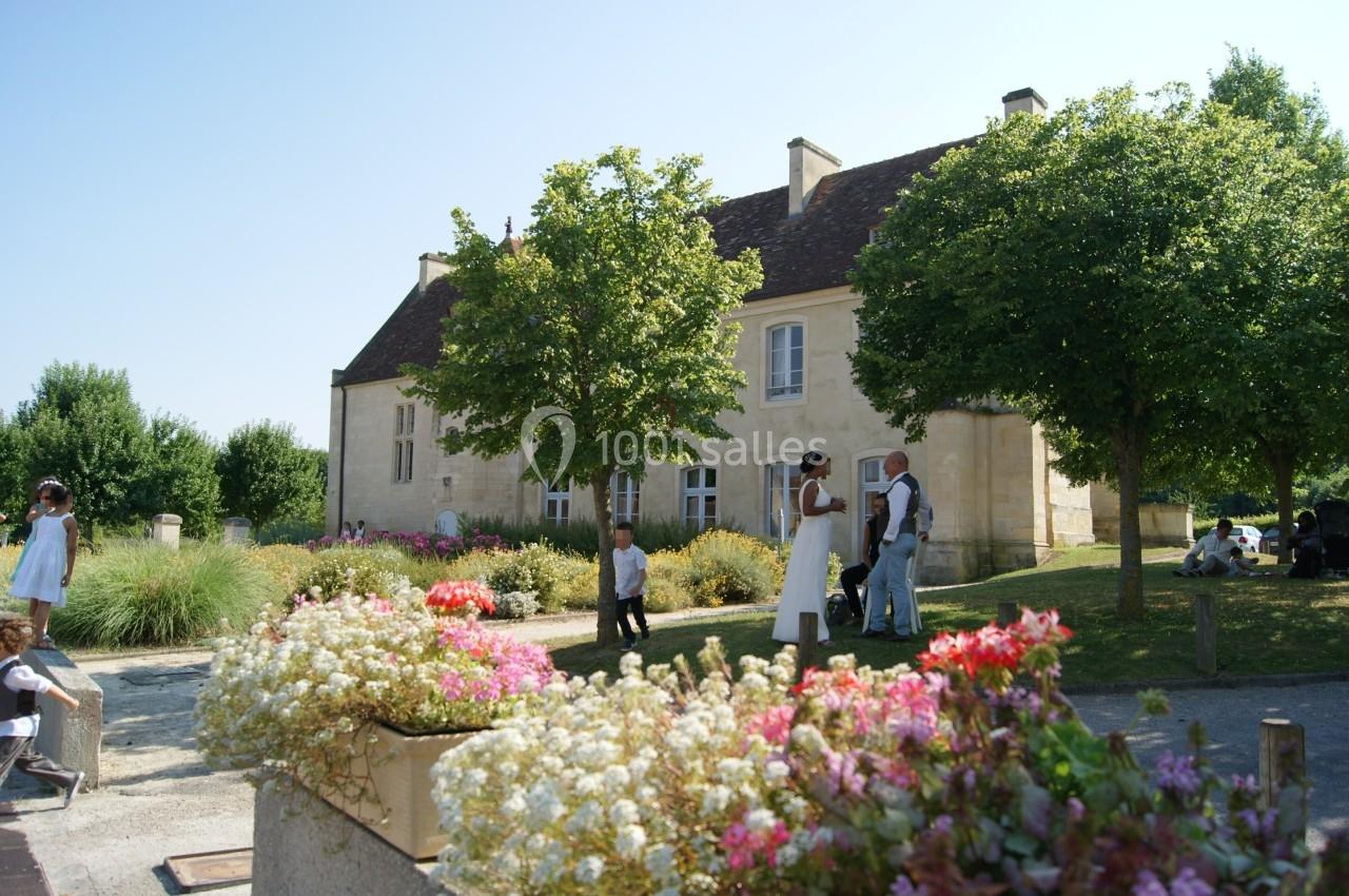 Des personnes discutent et se promènent dans un jardin fleuri devant un bâtiment en pierre sous un ciel dégagé.