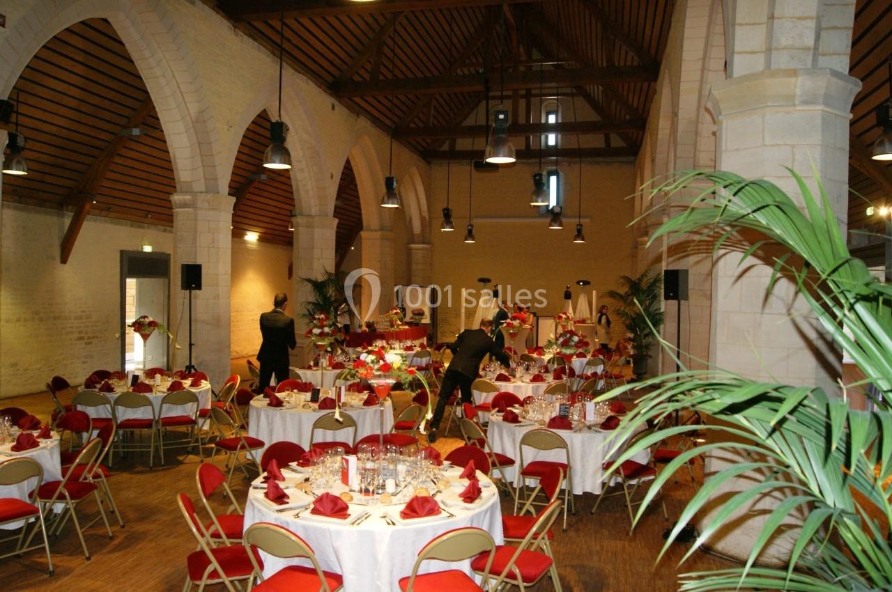 Salle de réception avec tables rondes dressées, nappes blanches, serviettes rouges et décoration florale.